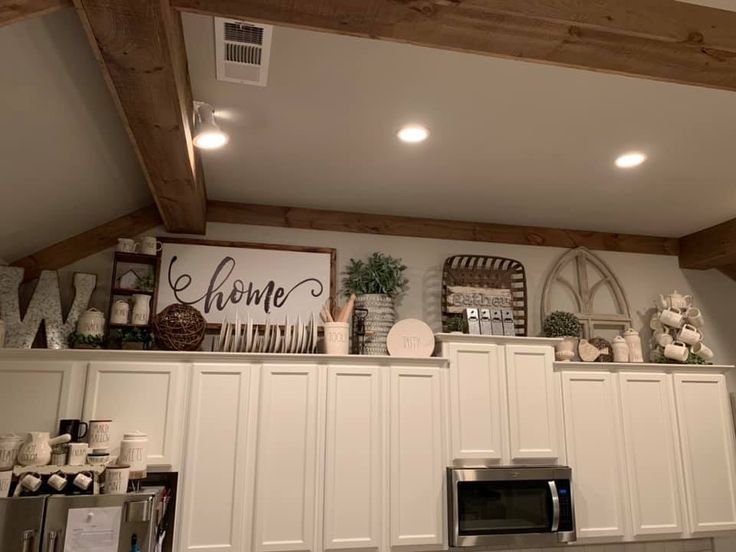 Collected above-cabinet display with a wood-framed home sign, a monogram letter, mugs, an arched mirror and a small potted plant above white kitchen cabinets