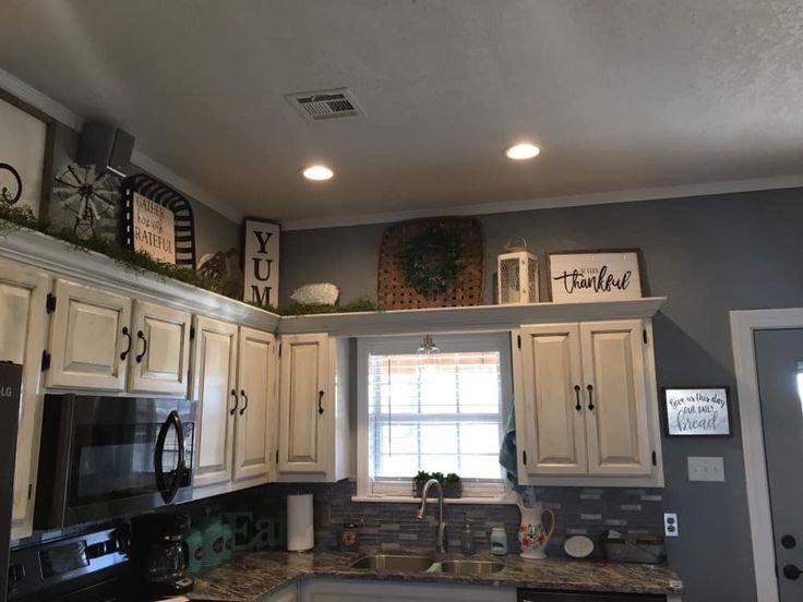 Framed signs, a woven wreath, a lantern and a windmill accent displayed above white kitchen cabinets near a window