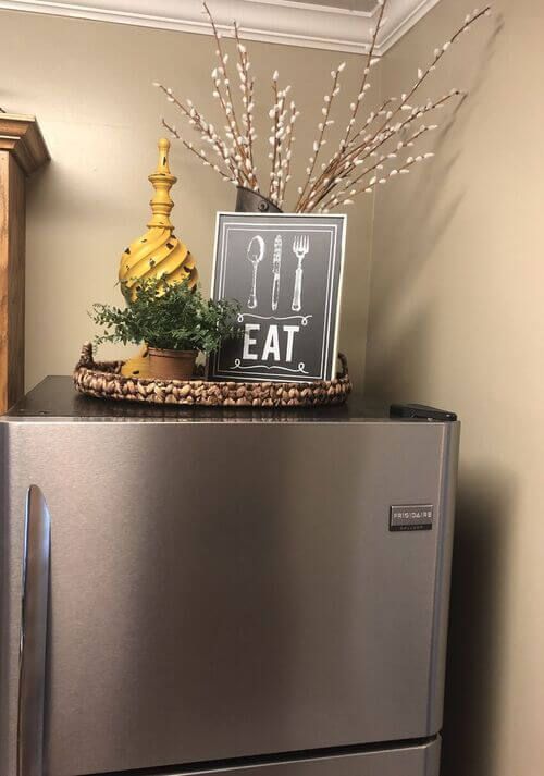 Small woven tray styled with an EAT sign and a decorative finial in a tight above-fridge space