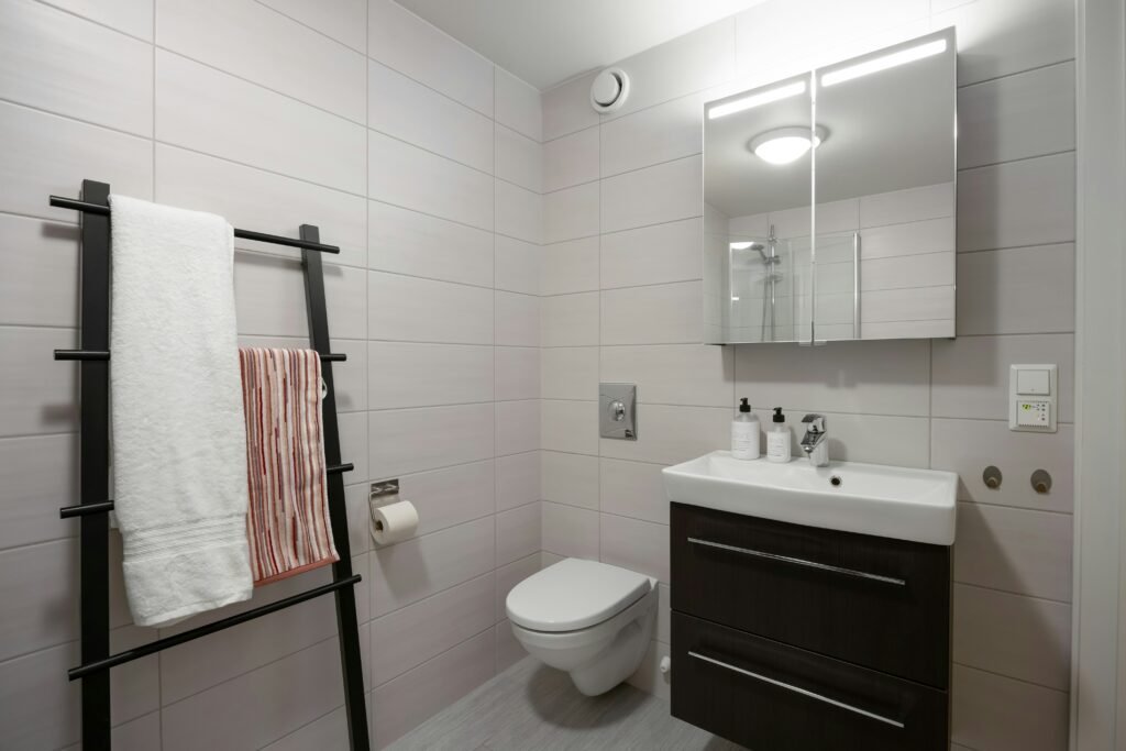 Backlit mirrored medicine cabinet above a dark wood vanity next to a black ladder towel rack holding a white towel and a striped towel
