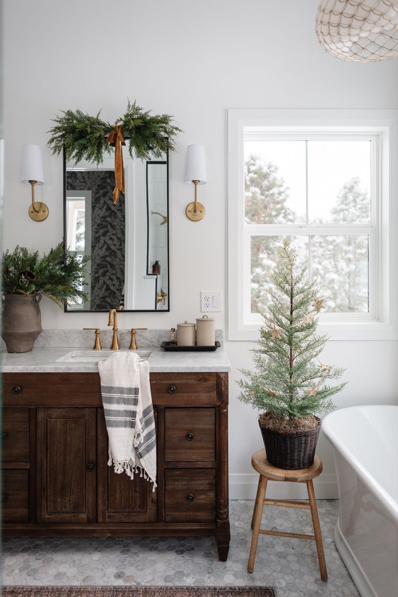 Garland-draped bathroom mirror flanked by brass sconces and a mini tree