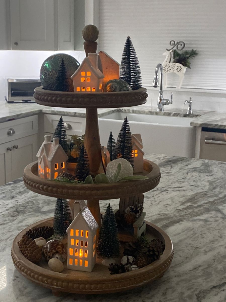Three-tier tray with lit white Christmas village houses and pinecones