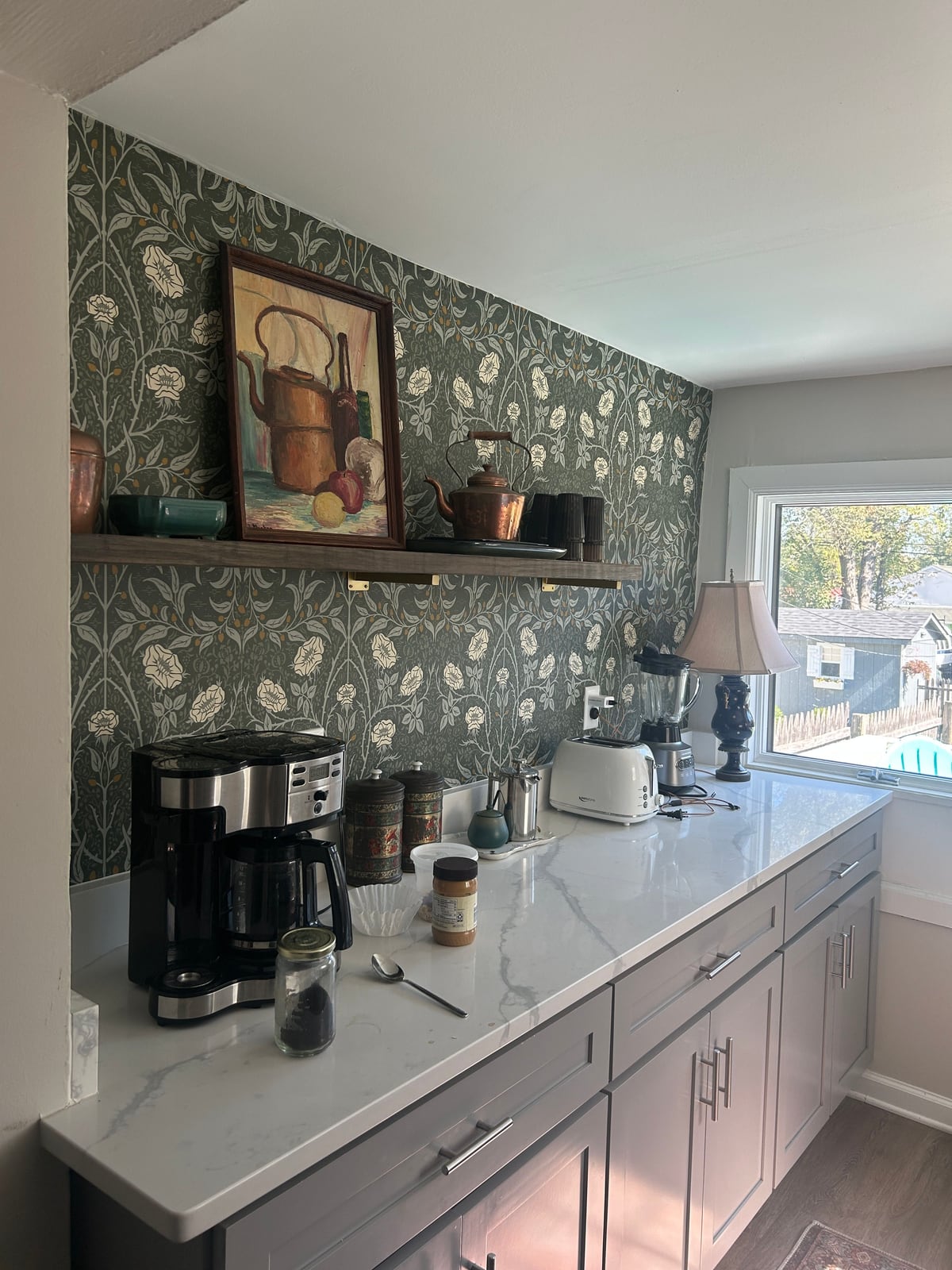 Kitchen counter with a coffee maker and toaster in front of green botanical wallpaper and a framed painting of a copper kettle