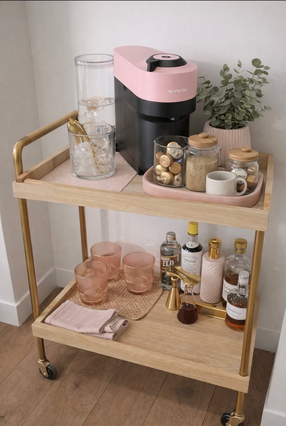 Two-tier wood and brass rolling cart with a pink coffee machine and glass jars on top and pink glasses and bottles on the lower shelf