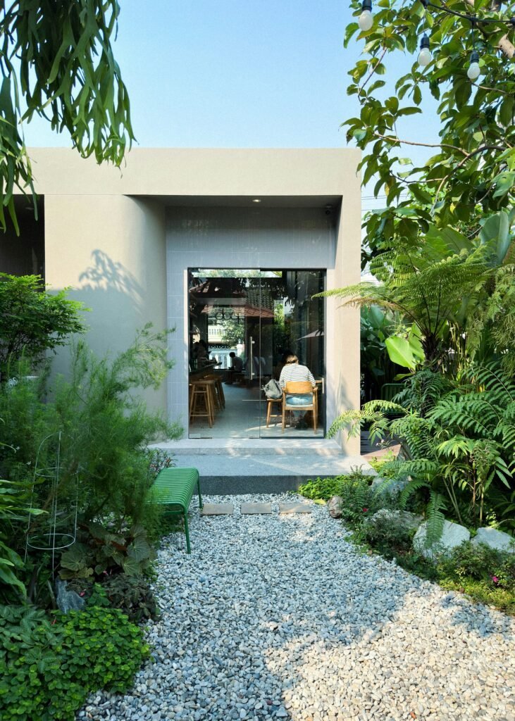 Light gravel garden pathway flanked by lush greenery and ferns leading toward a glass entryway