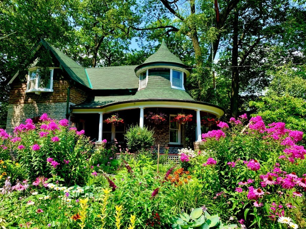 Victorian cottage with a turret and wraparound porch framed by dense pink and purple flower borders in full summer bloom