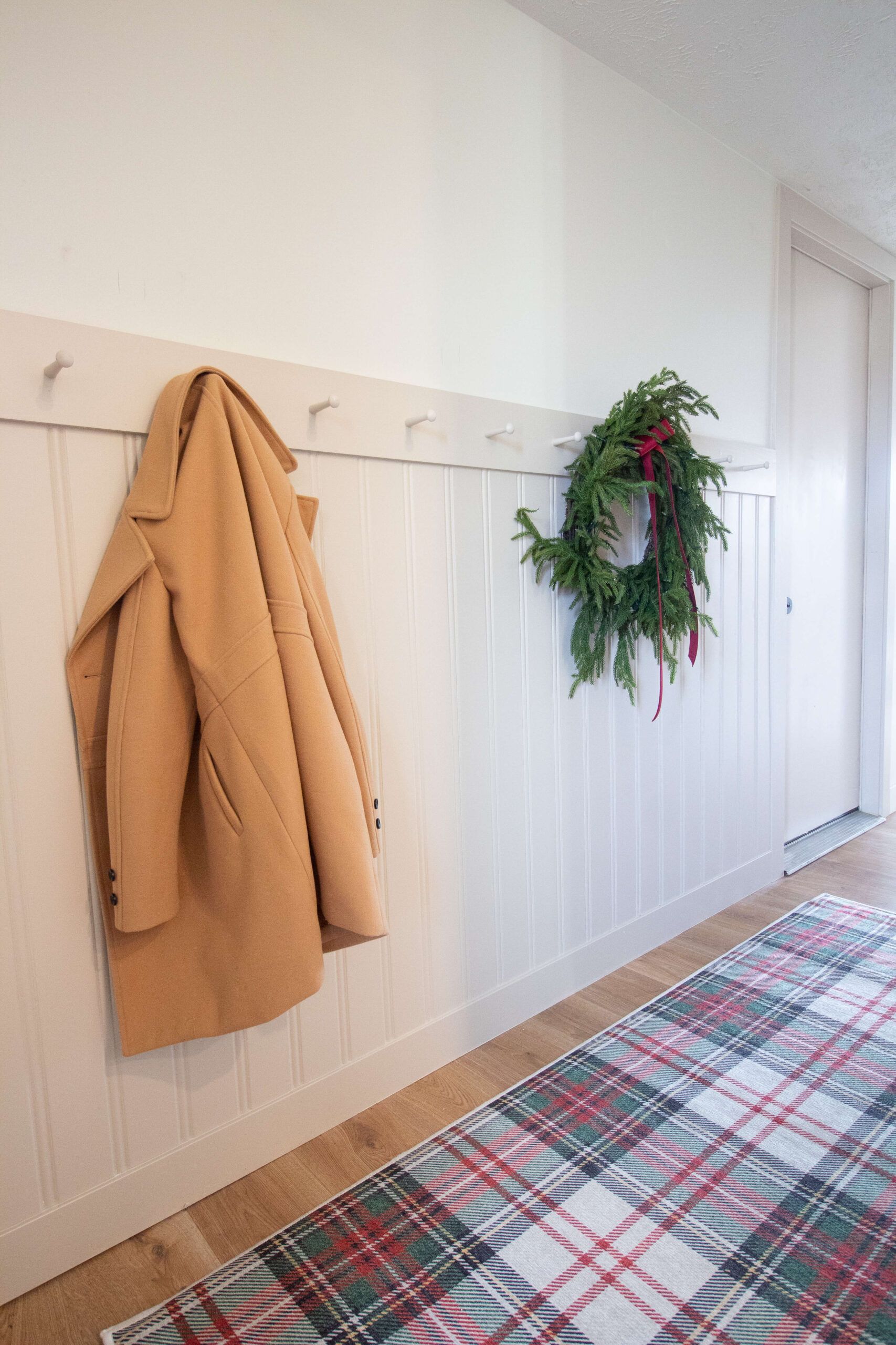 A beadboard hallway wall with a white peg rail, a camel coat and an evergreen wreath