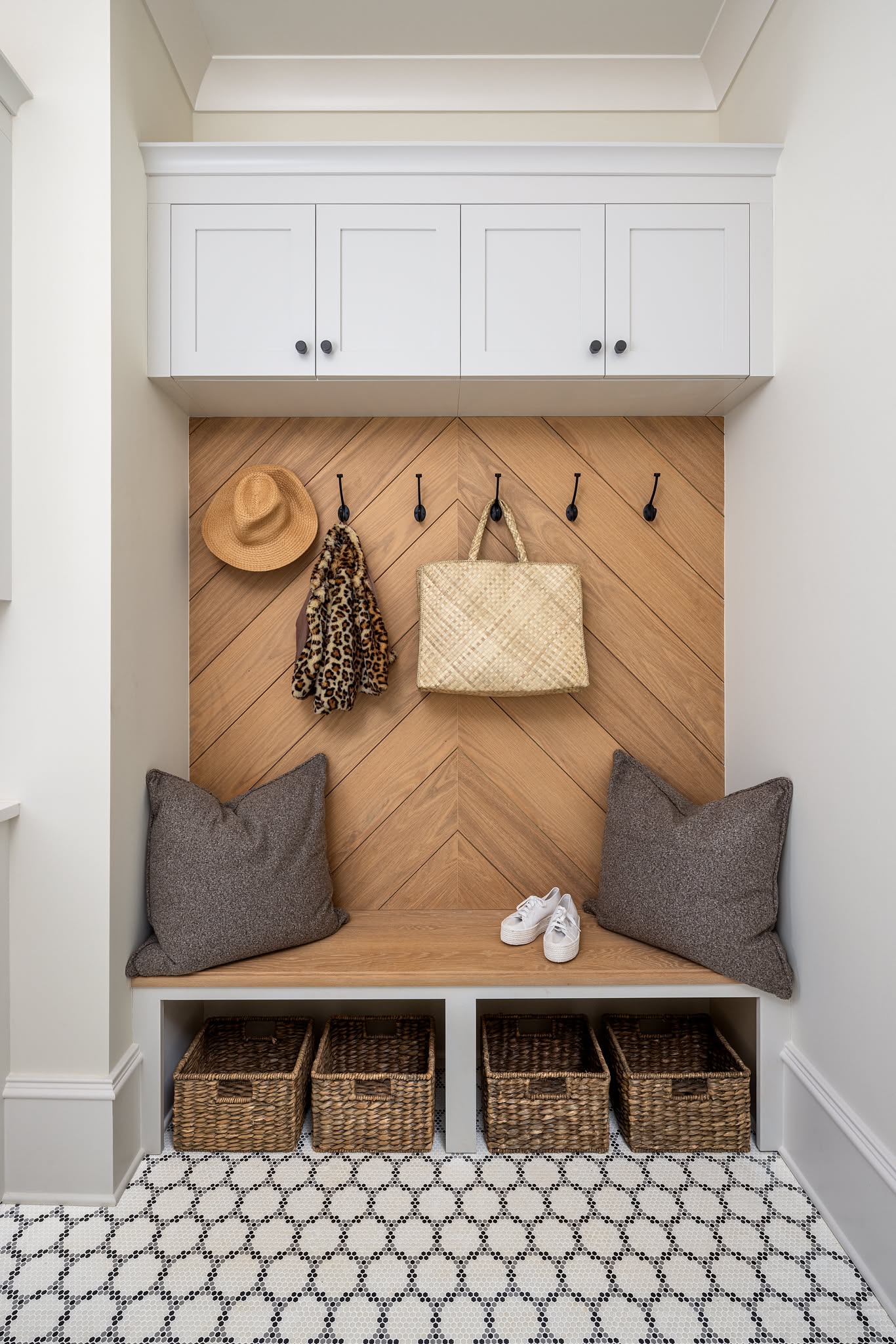 A herringbone wood paneled mudroom niche with white upper cabinets, a bench and four baskets