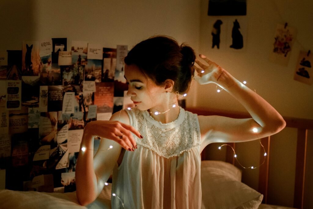 Fairy lights draped around a person's arms in a dorm room with a dense photo and postcard collage covering the wall behind them