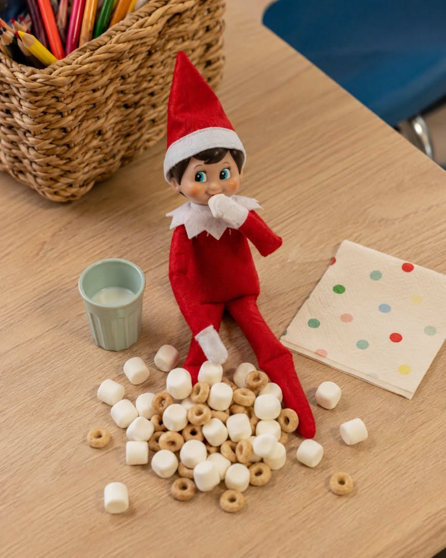 Elf on the Shelf sitting on a desk next to a pile of scattered cereal and mini marshmallows, with a cup of milk and a polka dot napkin