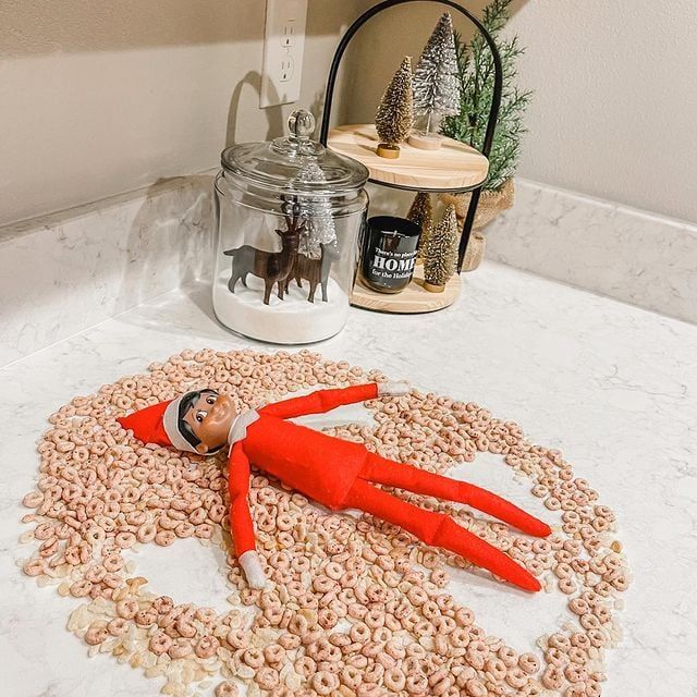 Elf on the Shelf lying in a pile of cereal shaped like a snow angel, next to a jar with a mini reindeer figurine