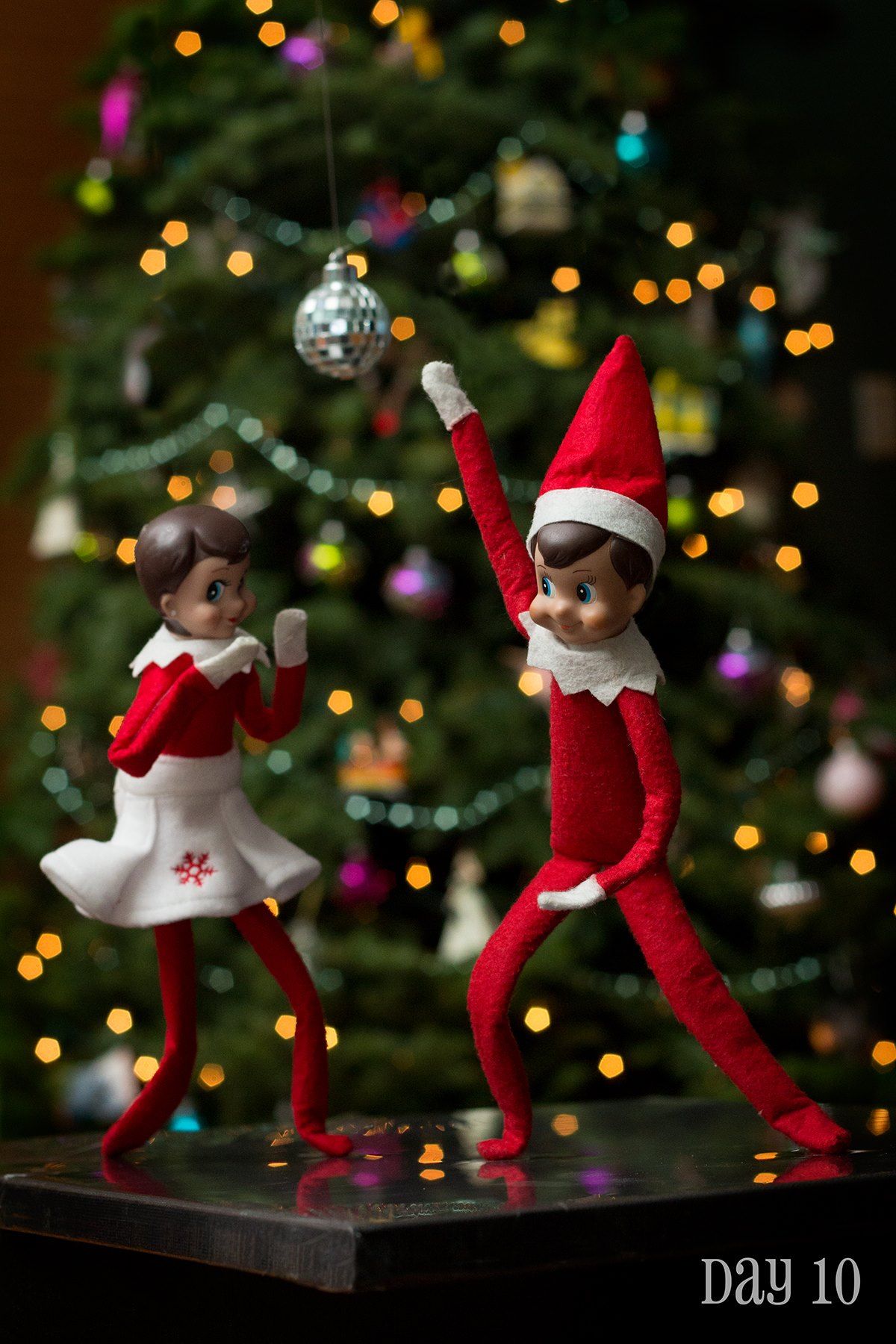 Two Elf on the Shelf dolls posed mid-dance move in front of a Christmas tree with a disco ball ornament hanging above them