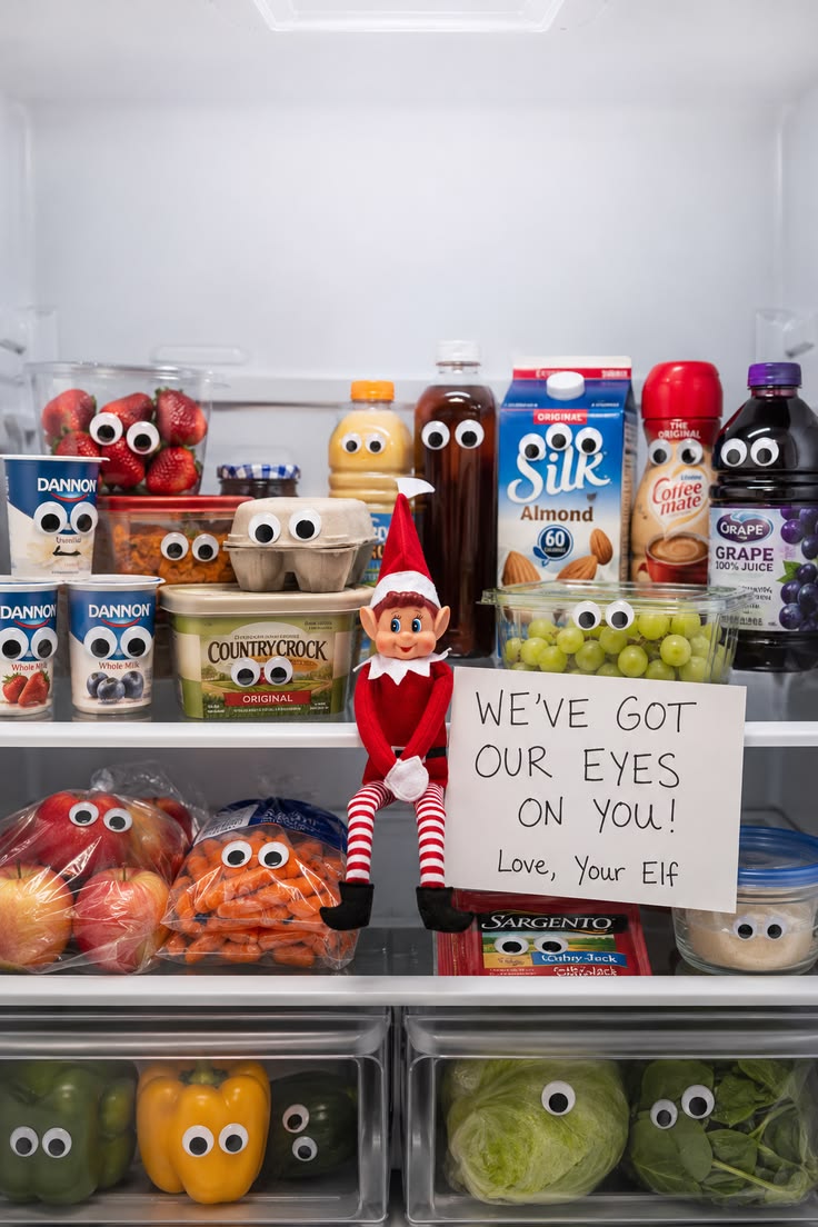 Refrigerator shelves full of food items with googly eyes stuck on them and a sign reading We've got our eyes on you, with the elf peeking out