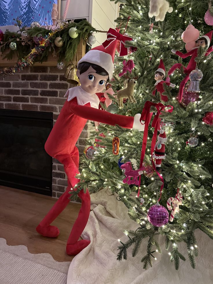 Large Elf on the Shelf figure standing beside a decorated Christmas tree, reaching into the branches as if hanging an ornament