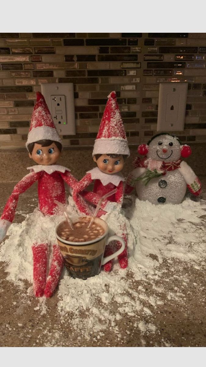 Two Elf on the Shelf dolls sitting in powdered snow next to a powdered-sugar snowman, sharing a mug of hot cocoa