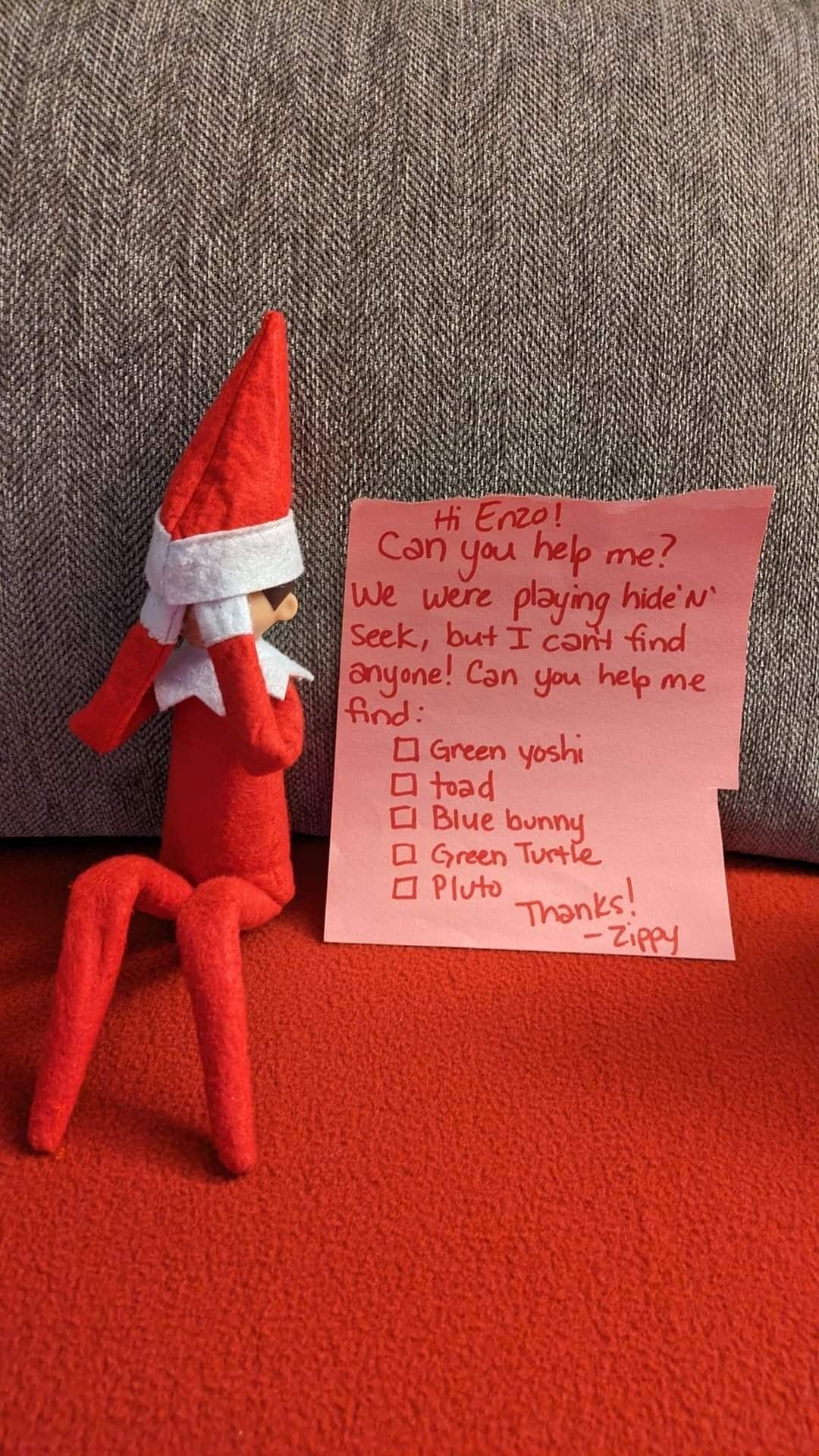 Elf on the Shelf sitting on the floor next to a handwritten checklist of toy characters to find, signed from the elf