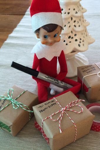 Elf on the Shelf holding a marker and writing gift tags on wrapped Christmas presents under the tree