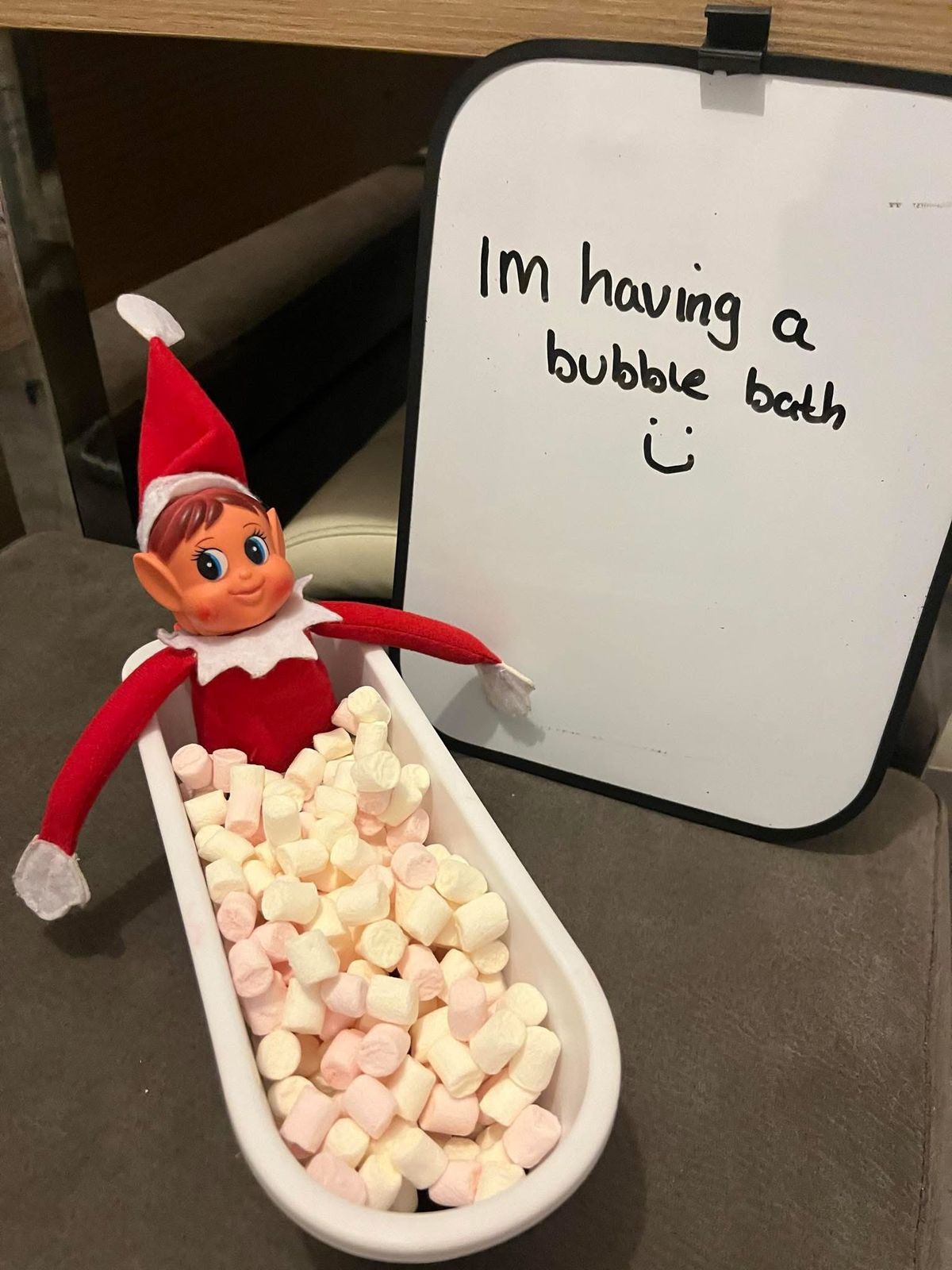 Elf on the Shelf sitting in a toy bathtub filled with marshmallows next to a whiteboard sign reading I'm having a bubble bath