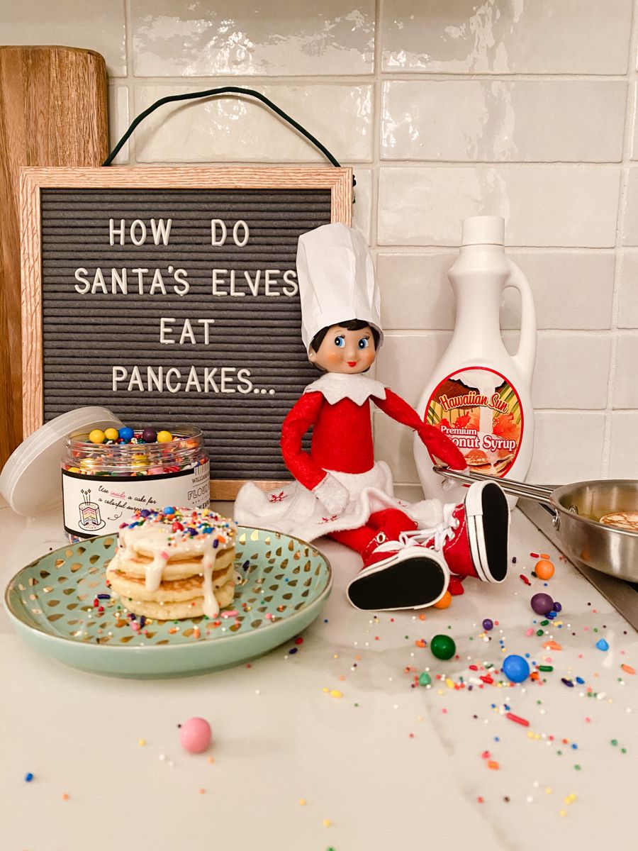 Elf on the Shelf wearing a chef hat beside a stack of pancakes topped with sprinkles and syrup, next to a letterboard sign