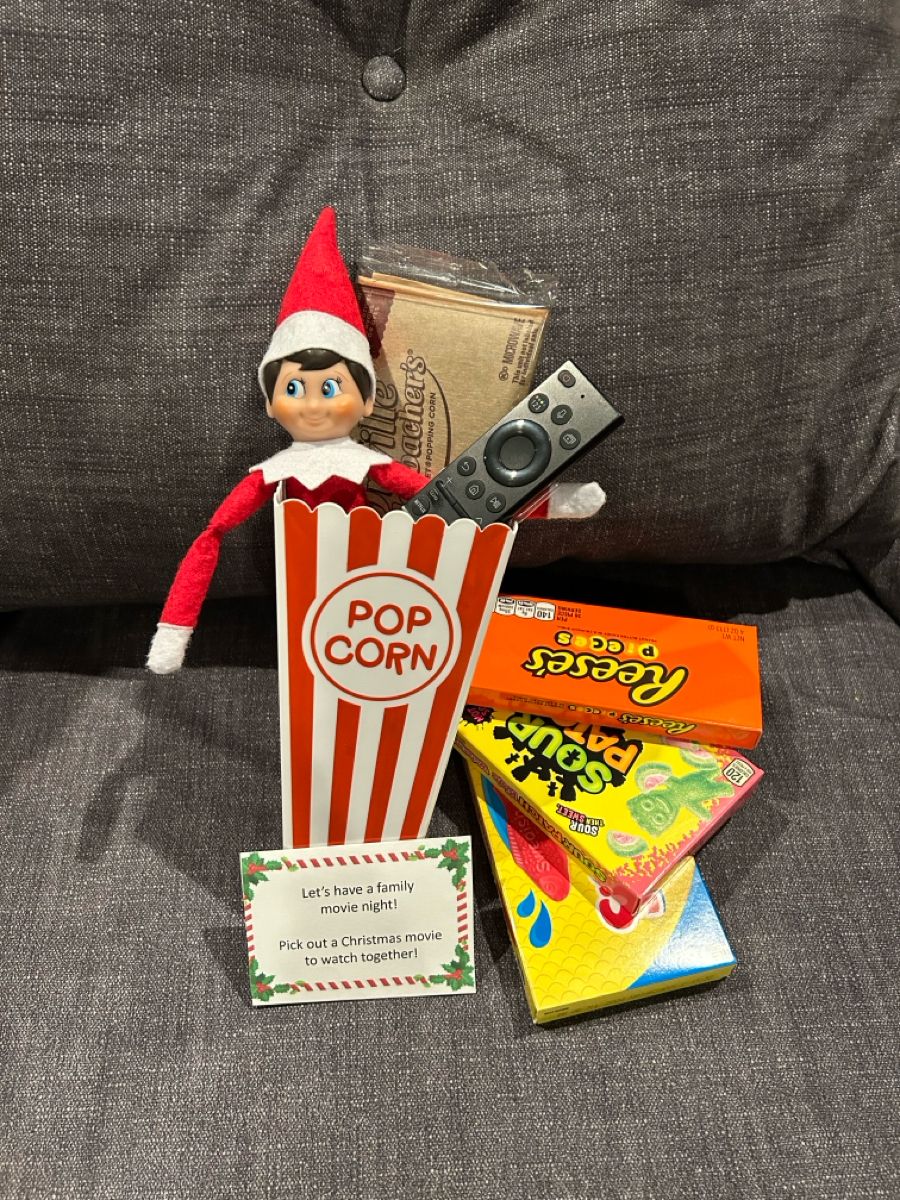 Elf on the Shelf sitting inside a popcorn box holding a TV remote, surrounded by candy boxes and a note that says let's have a family movie night