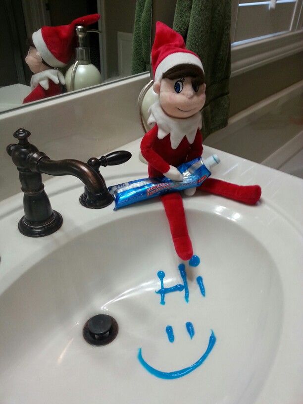 Elf on the Shelf sitting on a bathroom sink beside a toothpaste smiley face doodle