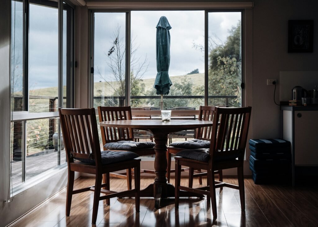 Round wood dining table with four chairs positioned by floor-to-ceiling windows overlooking rolling hills