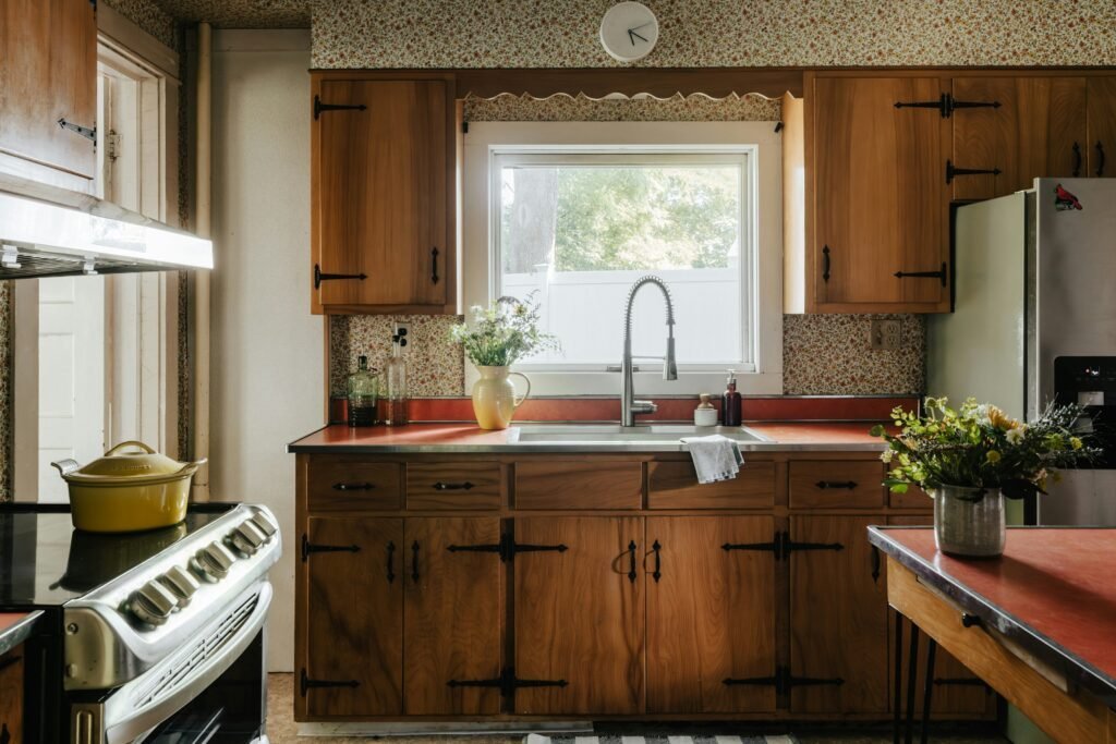 Vintage wood kitchen cabinets with wrought iron hinges and floral wallpaper beside a window with a red countertop