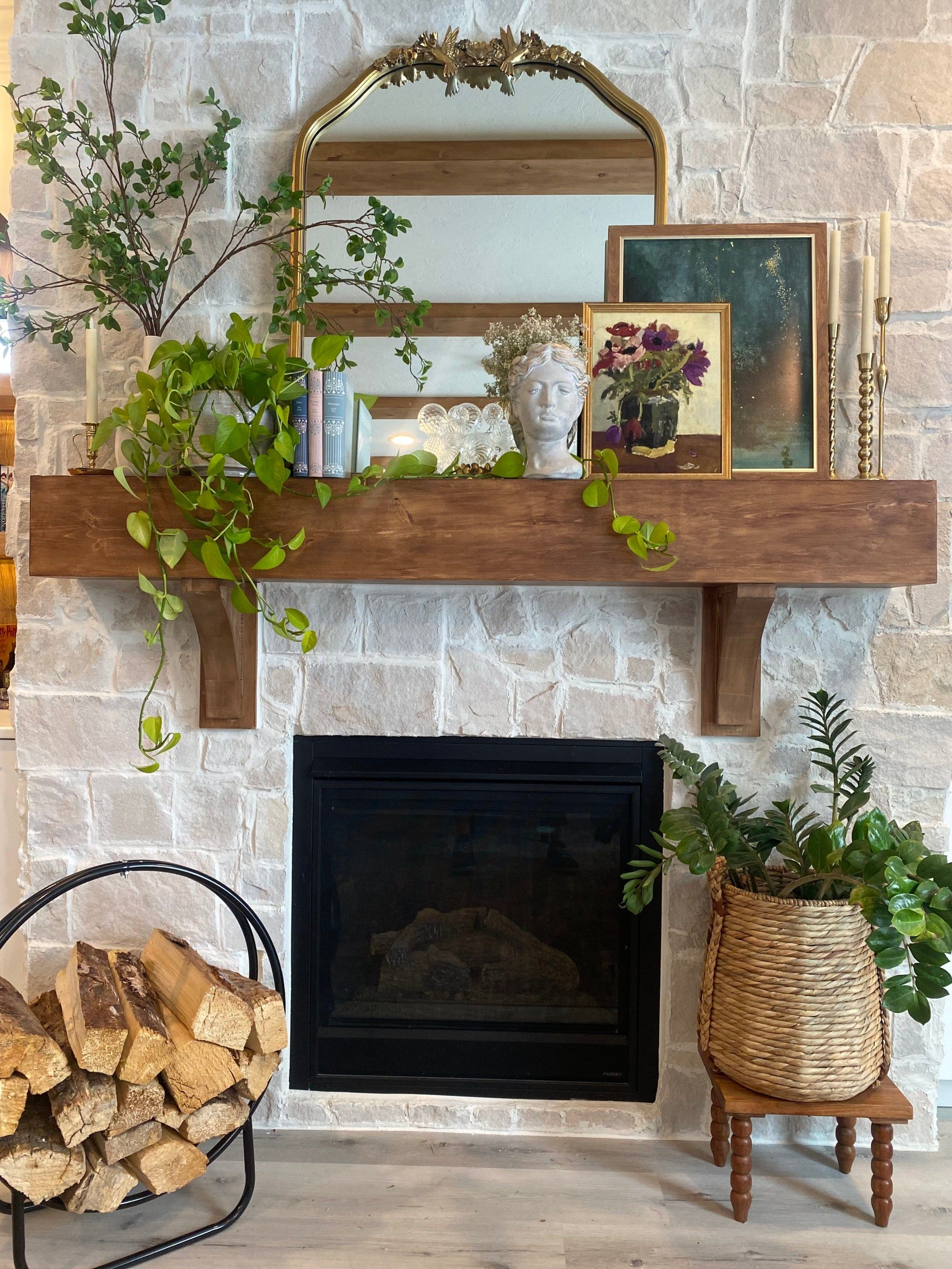 Eclectic mantel with a sculptural bust, layered framed paintings, trailing plants, brass candlesticks and an ornate gold mirror against a stone fireplace