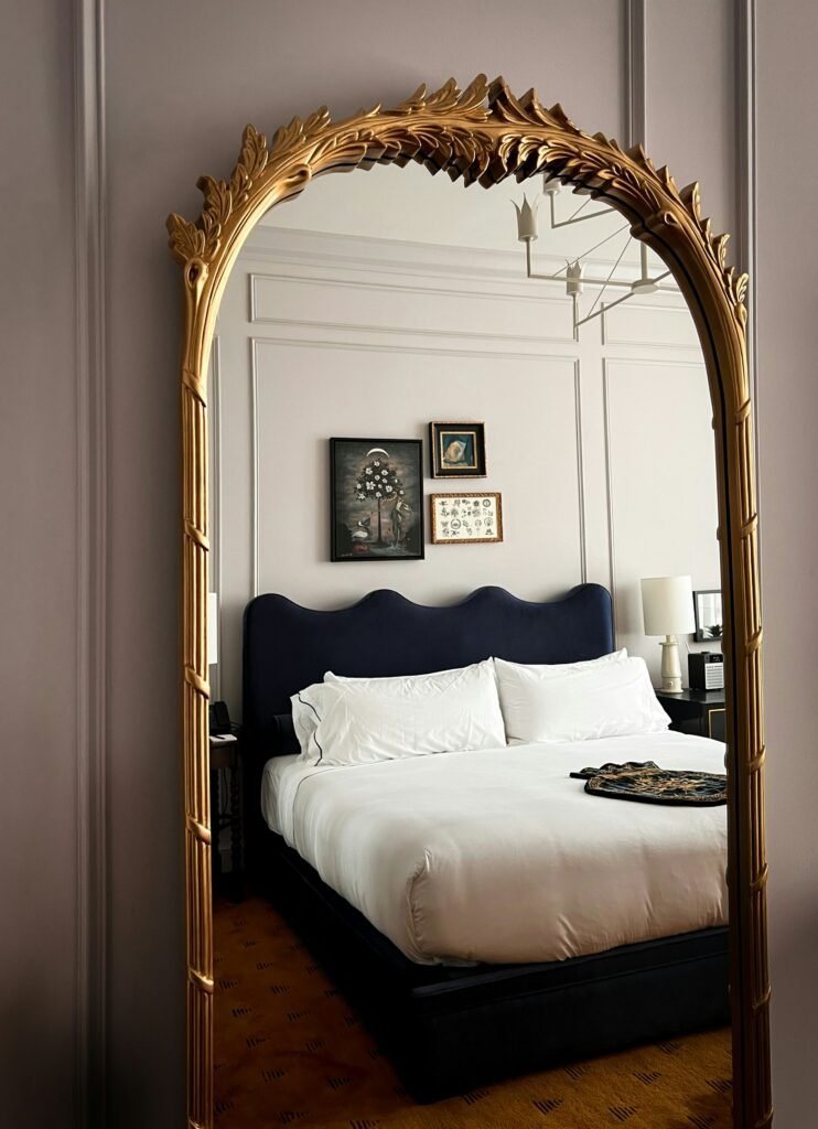 Ornate gold-carved arched mirror reflecting a bedroom with a navy upholstered headboard
