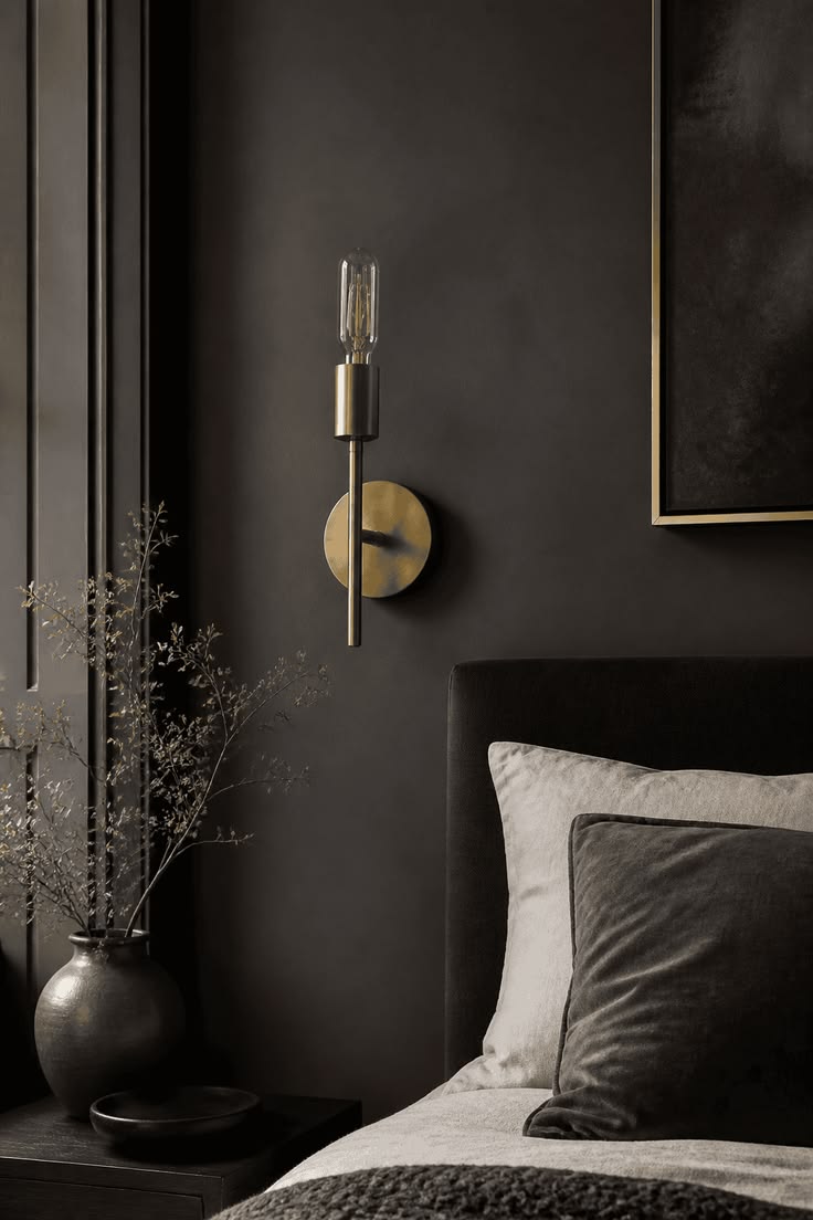 Cream and soft gray linen pillows glowing against a near-black wall with a dark velvet headboard and brass sconce
