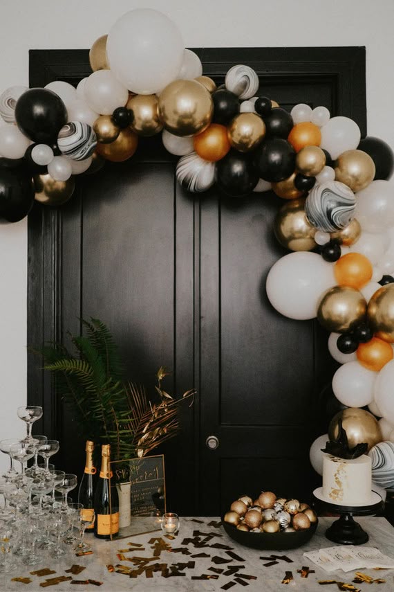 A black, gold, and marble balloon arch over a dark doorway with champagne and a cake nearby