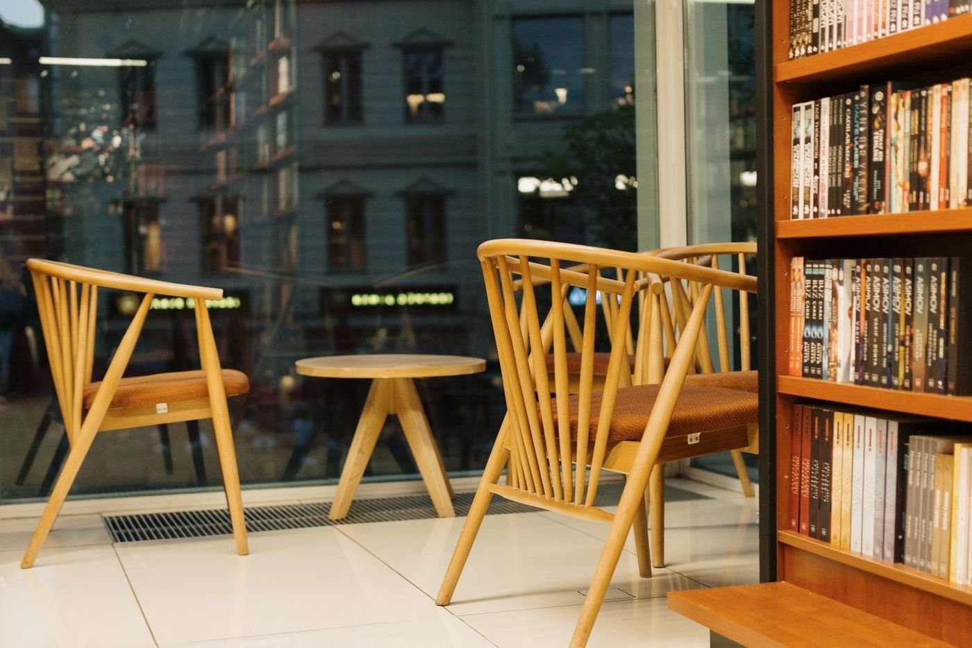 Two mid-century wood chairs with a small round table beside a window, positioned next to a tall bookshelf in a warmly lit room