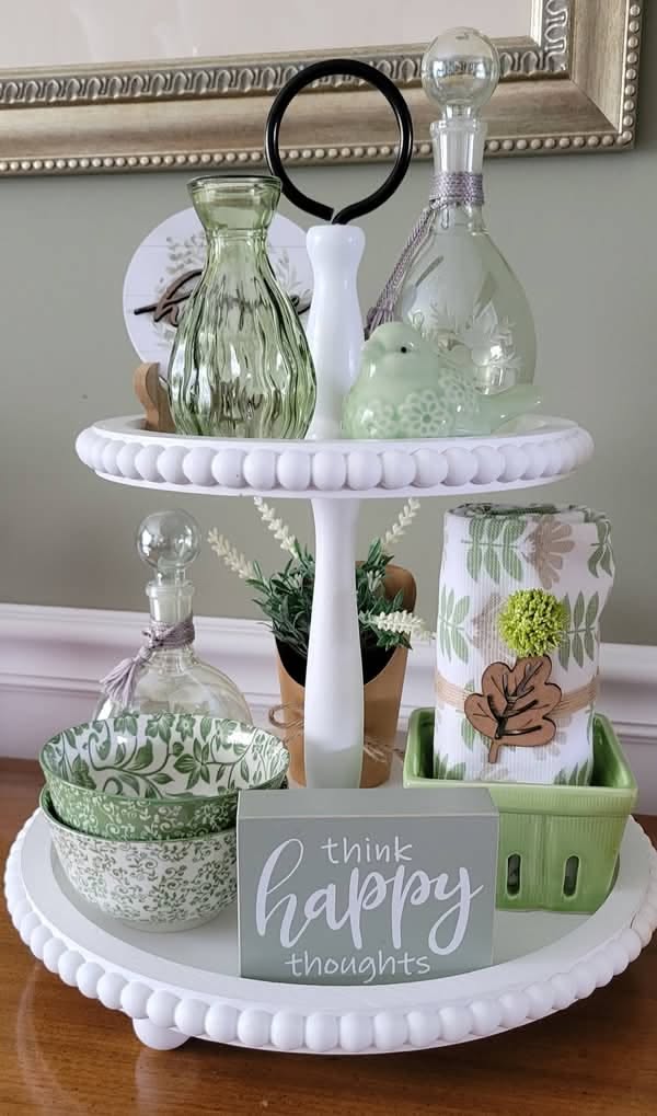 White two-tier tray styled simply with green glass bottles, a ceramic bird, patterned bowls, a rolled floral towel and a think happy thoughts sign