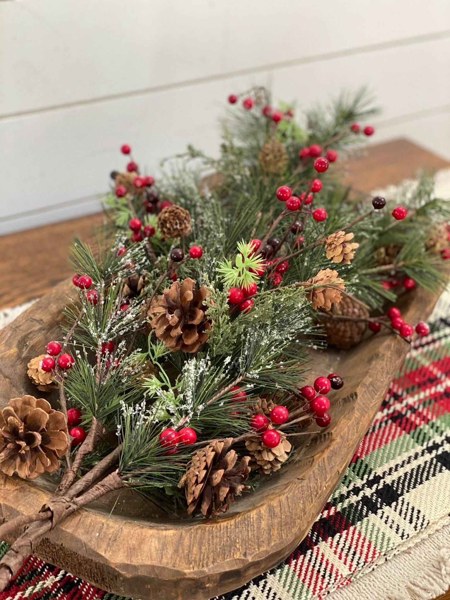 Dough bowl overflowing with frosted pine branches, red winter berries and pinecones on a plaid table runner
