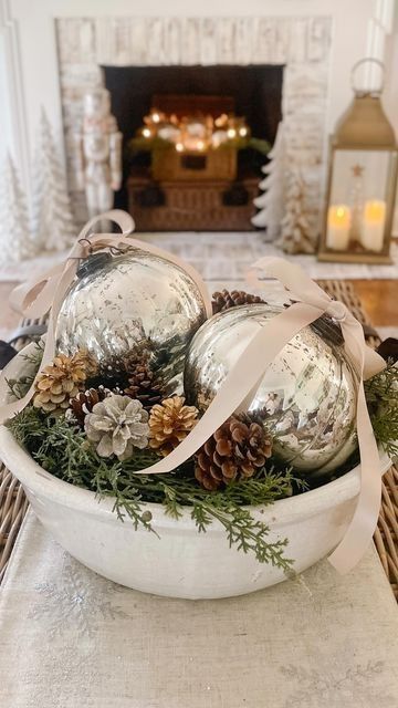 White ceramic bowl with two oversized mercury glass ornaments tied with ribbon, pinecones and greenery in front of a fireplace