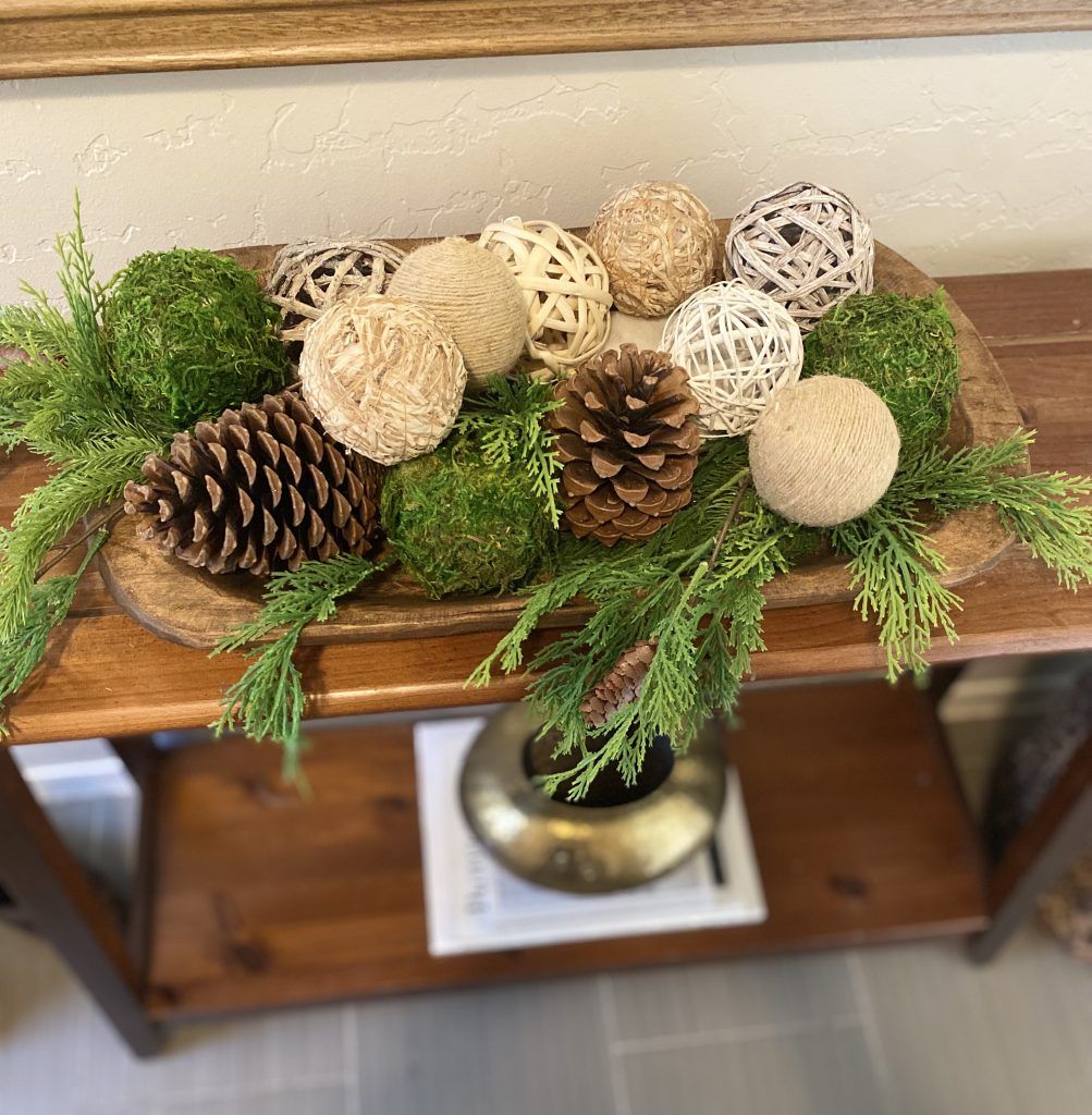 Dough bowl filled with moss balls, twine and rattan balls, pinecones and cedar branches on a console table