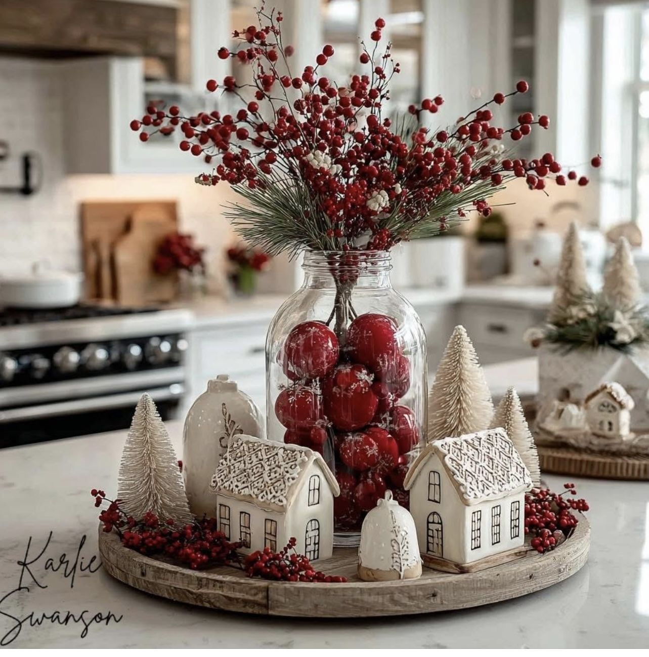 Round wood tray with a jar of red ornaments topped with berry stems, white ceramic houses and soft trees on a kitchen island