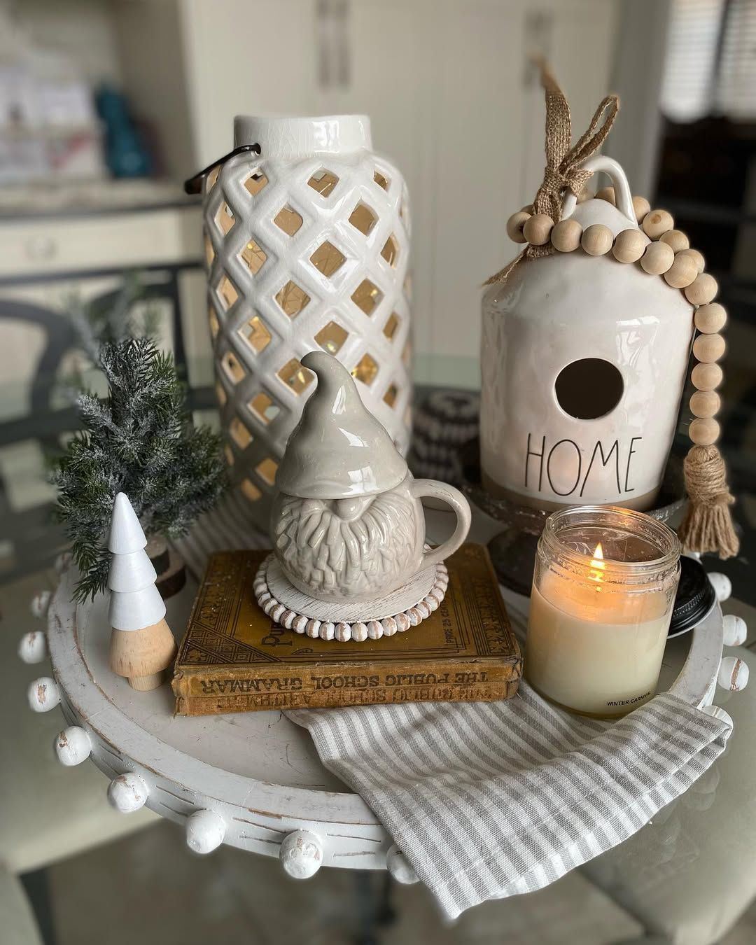 White beaded tray with a lattice lantern, a HOME birdhouse with wood beads, a knit gnome mug, a small tree and a candle
